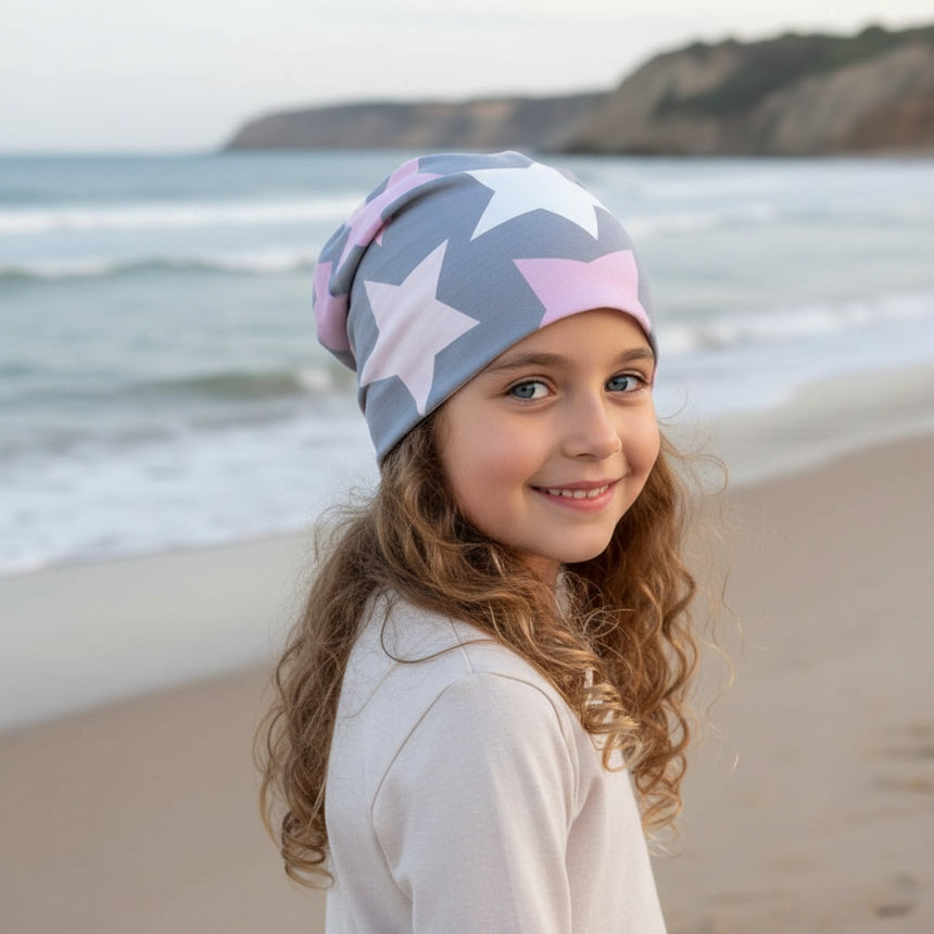 Ein Mädchen trägt die Wollhuhn Öko Long Beanie/Wendemütze mit großen Sternen, ideal für den lässigen Look am Strand. Handgemacht aus Öko-Jersey.