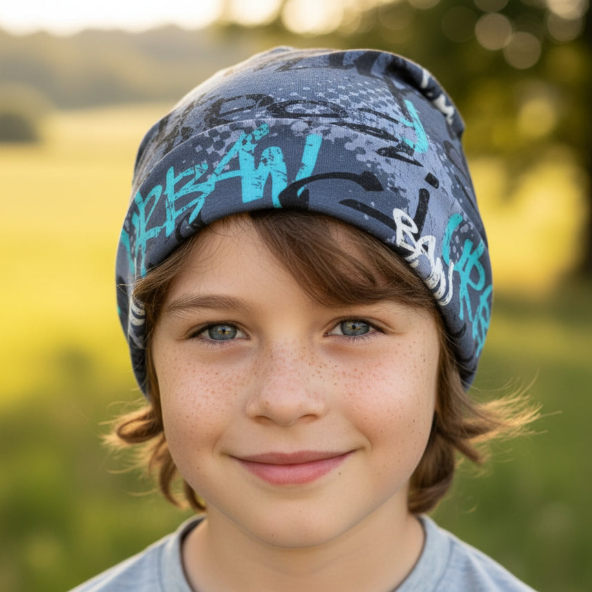 Junger Junge trägt Wollhuhn Öko Long Beanie, zweilagig, mit blauem Skater-Design. Handgefertigt aus weichem Jersey, ideal für einen lässigen, bequemen Look.