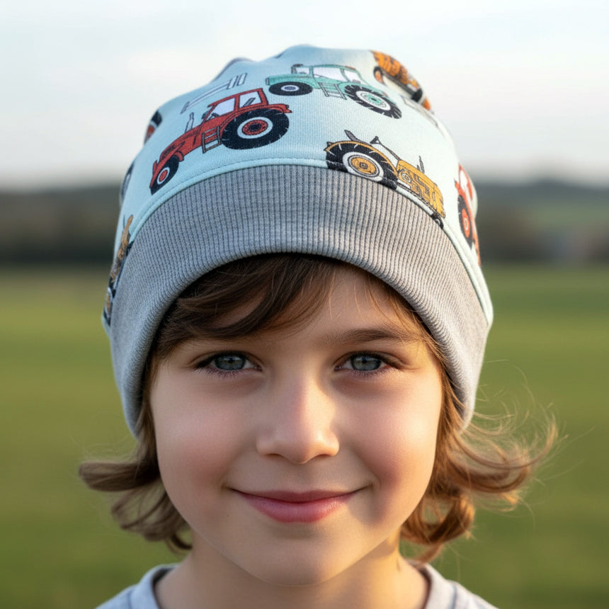Kind trägt Wollhuhn Dünne Öko Beanie-Mütze Sommer/Übergang, hellgrau mit bunten Traktor-Illustrationen, lächelt in einer Wiese. Handgemacht, bequem und ideal für Kinder.