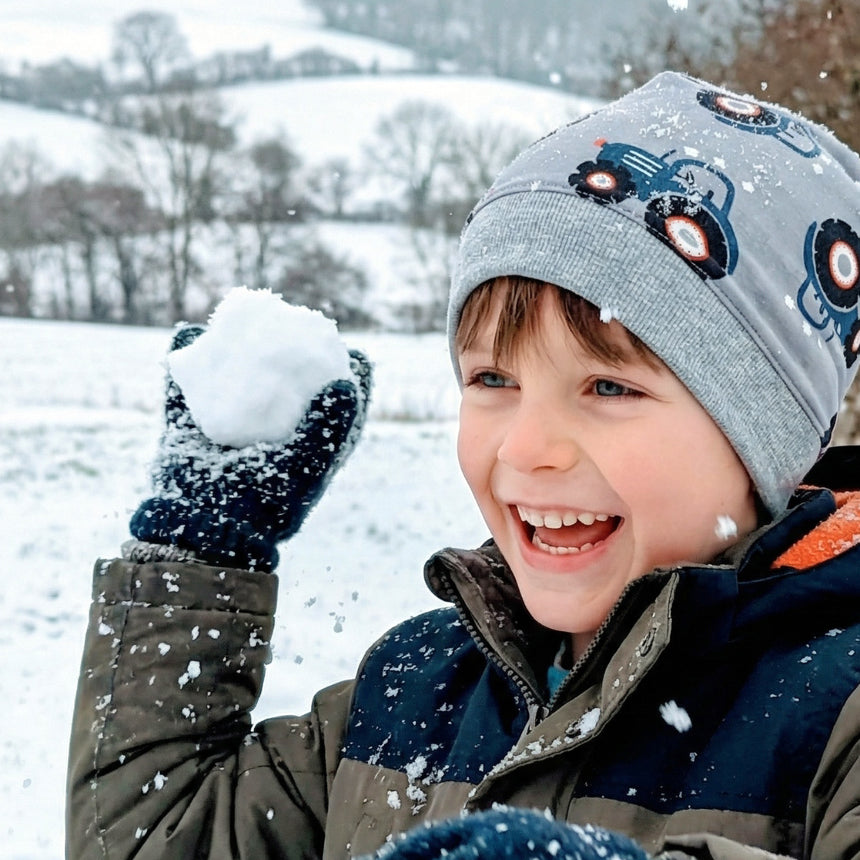 Junge mit Wollhuhn Dünne Öko Beanie-Mütze Maxi Traktor, grinsend im Schnee, hält Schneeball. Handgemacht, komfortabel und perfekt für aktive Kinder.