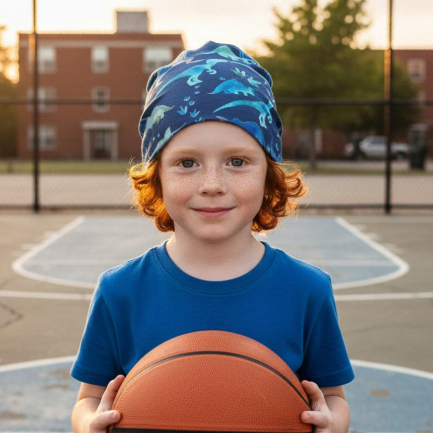 Kind mit rotem Haar trägt eine Wollhuhn Öko Long Beanie / Wendemütze, Ganzjährig, Zweilagig, Dinos Blau, auf einem Basketballplatz, hält Basketball.