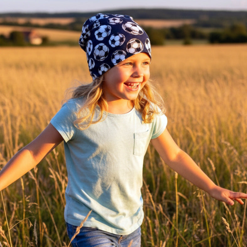 Ein Mädchen mit blonden Haaren trägt die Wollhuhn Öko Long Beanie mit Fußballmuster, spielt fröhlich in einem goldenen Feld bei Sonnenuntergang.