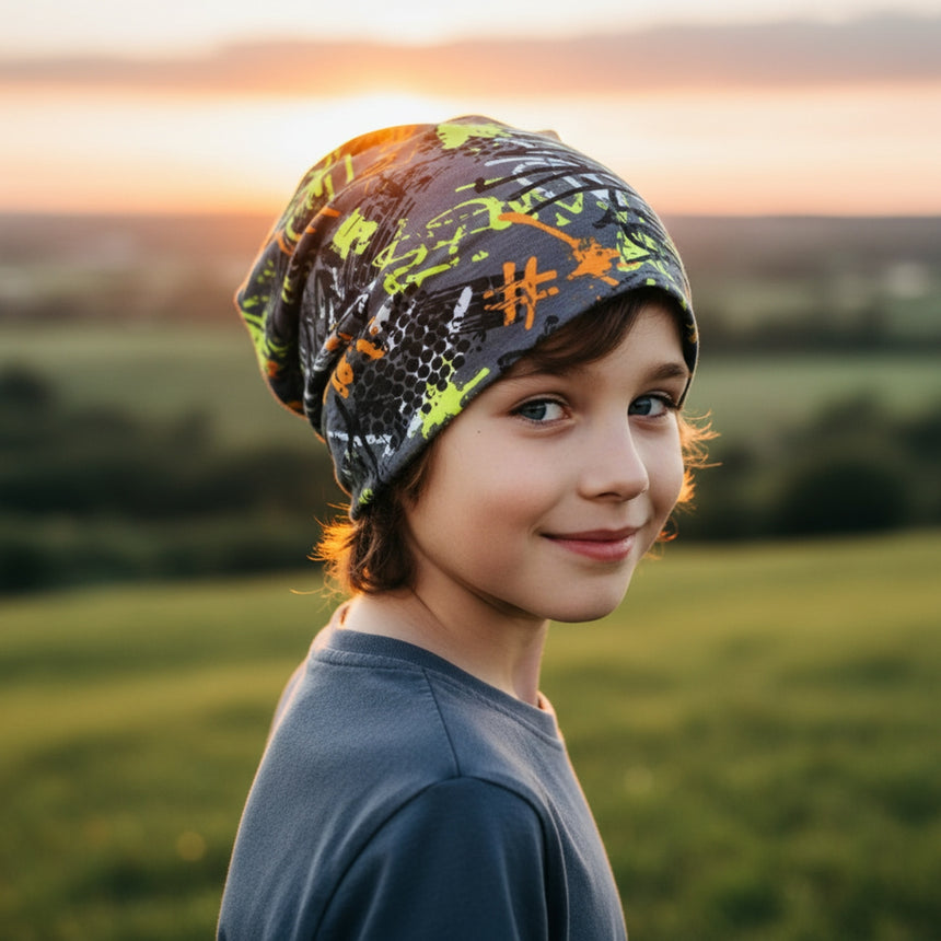 Junge mit Wollhuhn Öko Long Beanie, Wendemütze in Graffiti-Design, lässig fallend, im Sonnenuntergang auf einer Wiese stehend, symbolisiert handgemachte Qualität und Komfort.