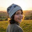 Kind mit Wollhuhn Dünne Öko Beanie-Mütze Sommer/Übergang Einlagig Maxi Traktor/Trecker Grau, lächelnd im Sonnenuntergang auf einer Wiese, trägt eine neutrale Jacke.