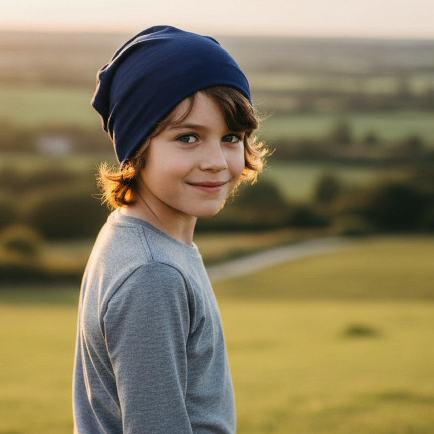 Ein Junge trägt eine Wollhuhn Öko Long Beanie in Dunkelblau, lässig am Hinterkopf fallend, während er in einer ländlichen Umgebung lächelt.