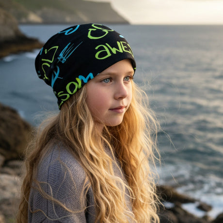 Mädchen mit Wollhuhn Öko Long Beanie, zweilagig, cooles Design Awesome, steht am felsigen Ufer, trägt einen grauen Pullover, Hintergrund: ruhiges Meer und Küste.