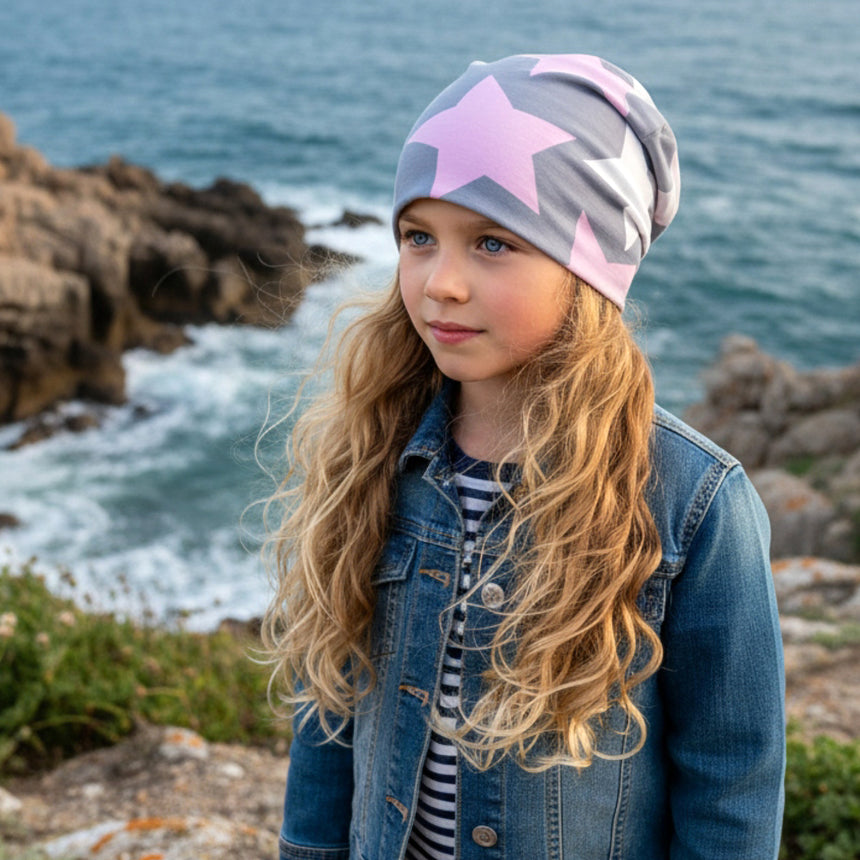 Kind mit Wollhuhn Öko Long Beanie, Wendemütze, ganzjährig tragbar, zweilagig, mit großen Sternen in Grau/Rosa/Pink, steht am felsigen Meeresufer.
