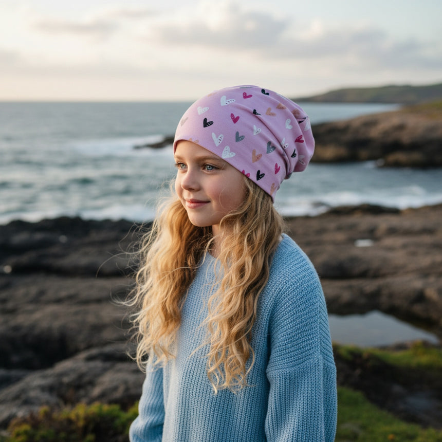 Mädchen am felsigen Ufer trägt Wollhuhn Öko Long Beanie mit Herzmuster, ideal für Kinder, zweilagig, wendbar, handgefertigt in Deutschland, ganzjähriger Komfort.