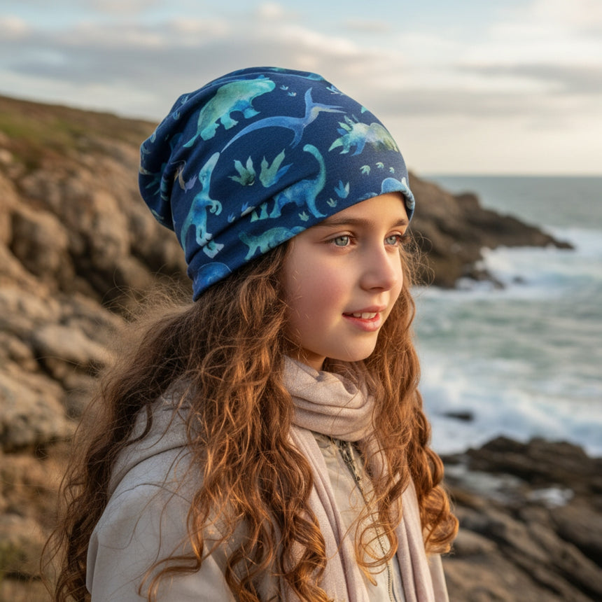 Ein Kind trägt eine blaue Wollhuhn Öko Long Beanie mit Dino-Muster am Meer, passend für Frühling bis milde Wintertage, handgemacht aus nachhaltigem Jersey.