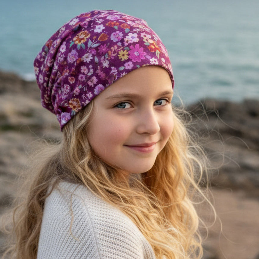 Mädchen mit blonden Haaren trägt eine Wollhuhn Öko Long Beanie / Wendemütze mit Blumenmuster, blickt lächelnd über die Schulter, Hintergrund: Felsenküste und Meer.