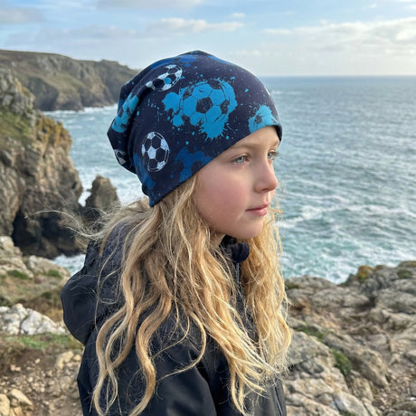 Kind mit blonden Haaren trägt Wollhuhn Öko Long Beanie, zweilagig, mit Fußballmotiven, ideal für das ganze Jahr. Steht am felsigen Küstenmeer.
