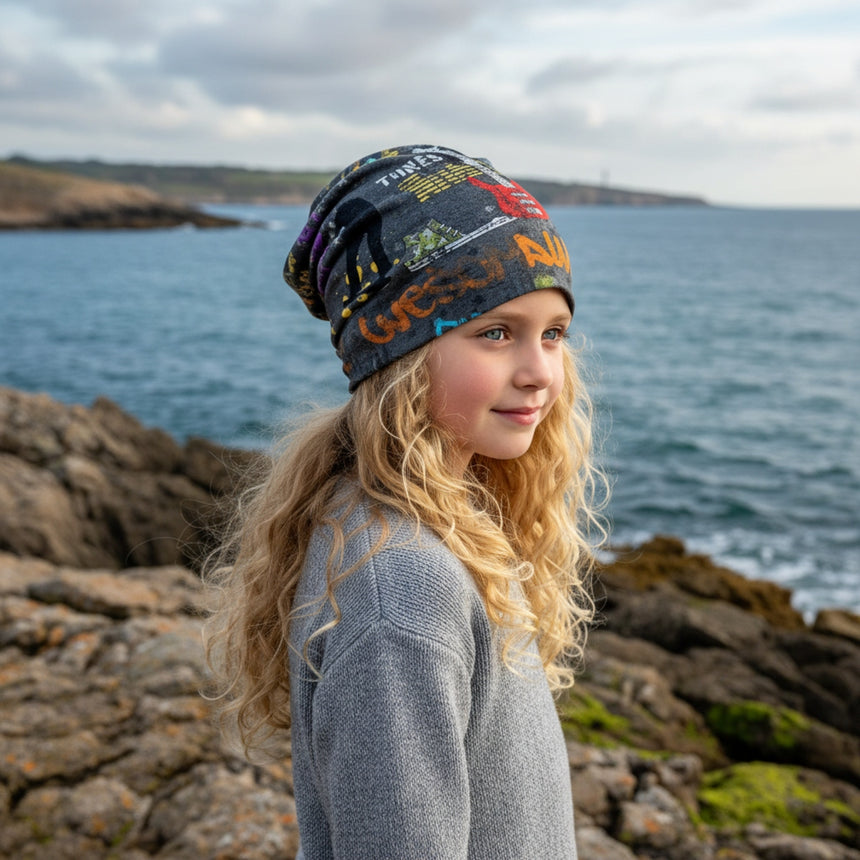 Ein Kind trägt die Wollhuhn Öko Long Beanie, eine zweilagige, wendbare Mütze mit coolen Graffiti-Motiven, ideal für lässigen Komfort am Meer.