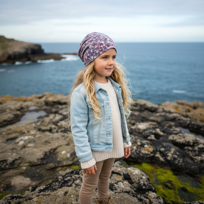 Junges Mädchen in Wollhuhn Öko Long Beanie in Blümchen Dark Pastell, an felsiger Küste stehend, trägt Denimjacke und Stiefel, symbolisiert handgefertigte Qualität und Komfort.