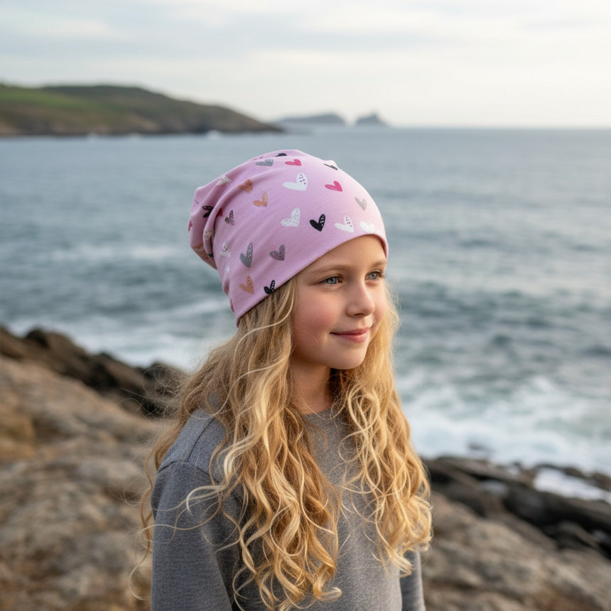 Mädchen mit blonder Wallemähne am Felsenstrand trägt Wollhuhn Öko Long Beanie, zweilagig, rosa Herzen, bequem und wendbar, ideal für das ganze Jahr.