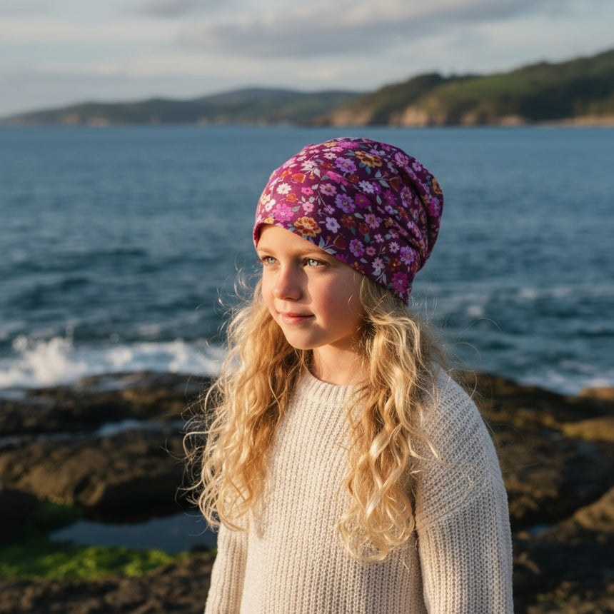 Mädchen am Meer trägt Wollhuhn Öko Shorties, Wendemütze Blumen Beere, handgenäht, weich und dehnbar, ideal für aktive Kinder, stilvoll und bequem.