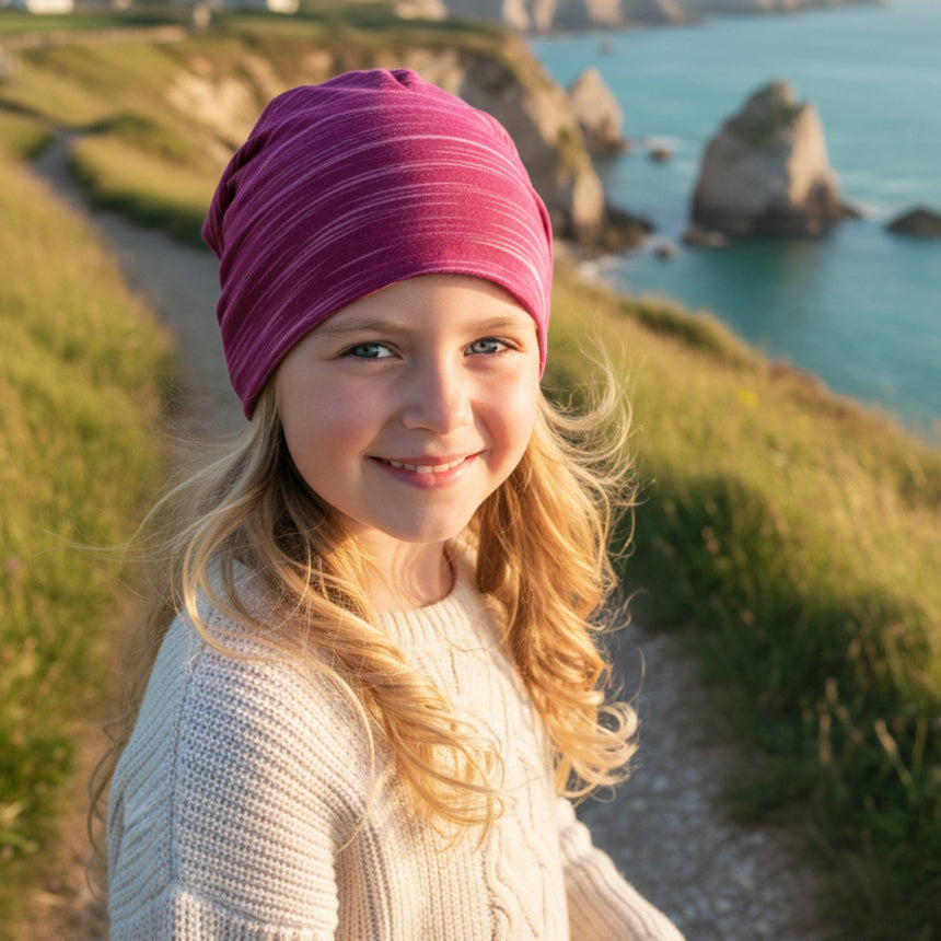 Mädchen auf Klippe trägt Wollhuhn Öko Shorties/Wendemütze, Ganzjährig, Zweilagig, Lines Beere, lächelt in Kamera, symbolisiert handgefertigte, nachhaltige Kinderaccessoires.