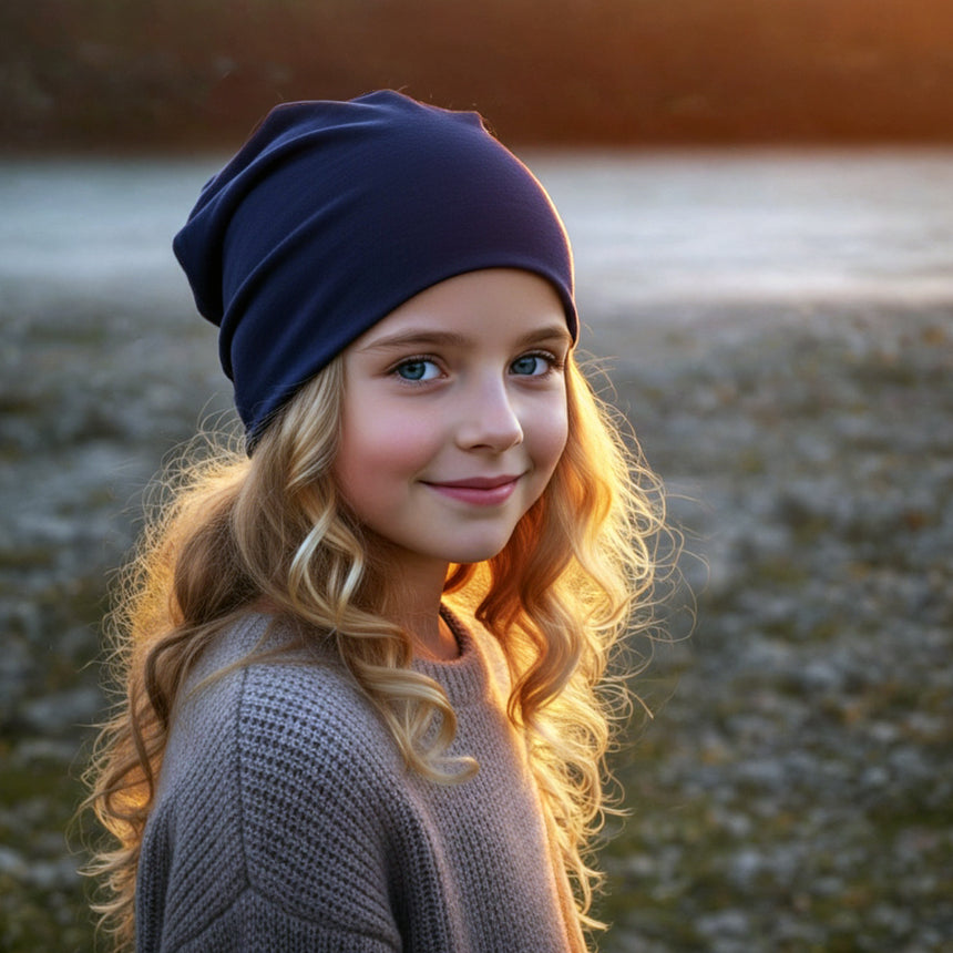 Mädchen am Flussufer trägt Wollhuhn Öko Shorties, dunkelblau. Handgemachte, zweilagige Wendemütze, ideal für aktive Kinder. Warm, dehnbar, passt unter Helme.