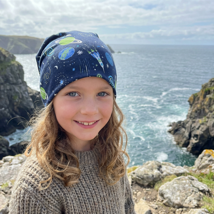 Kind auf Felsen am Meer mit Wollhuhn Öko Long Beanie, Wendemütze, zweilagig, Space/Rakete Dunkelblau, lächelt in die Kamera, handgefertigt aus weichem Öko-Jersey.