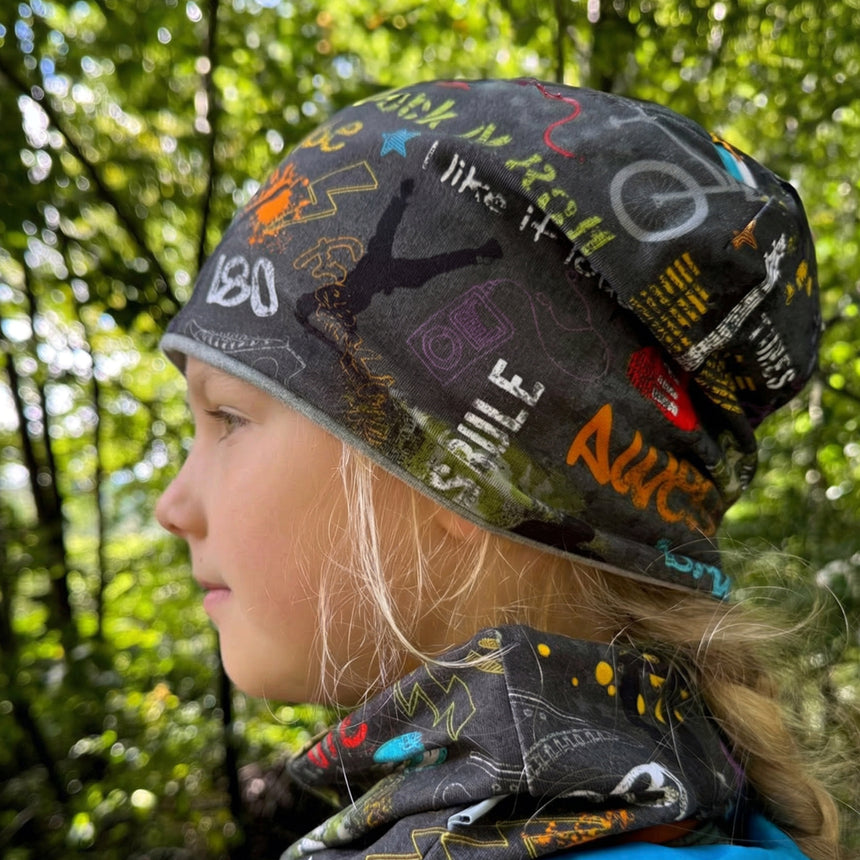 Ein Kind trägt eine Wollhuhn Öko Shorties Wendemütze mit buntem Graffiti-Muster im Wald, ideal für aktive Kinder, handgemacht und zweilagig.