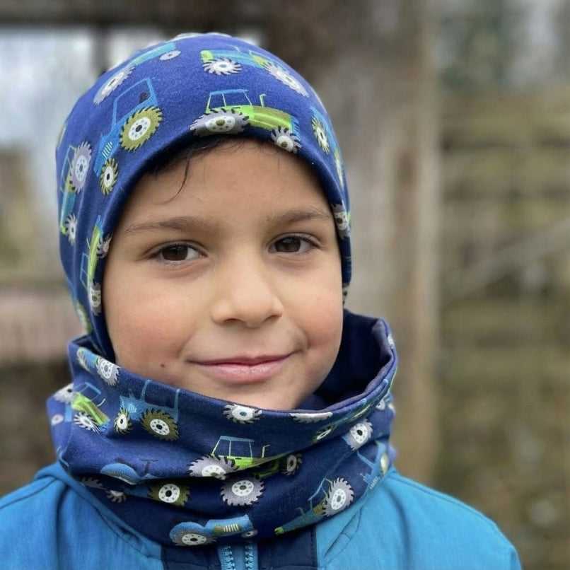 Wollhuhn Öko Shorties: Kind im blauen Beanie mit Traktormuster, lächelnd im Freien, passend für aktive Kinder, handgemacht aus weichem Öko-Jersey, zweilagig, wendbar.