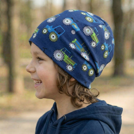 Kind mit Wollhuhn Öko Shorties Wendemütze, dunkelblau mit Traktormotiven, lächelt im Profil. Handgemachte, zweilagige Beanie, ideal für aktive Kinder.