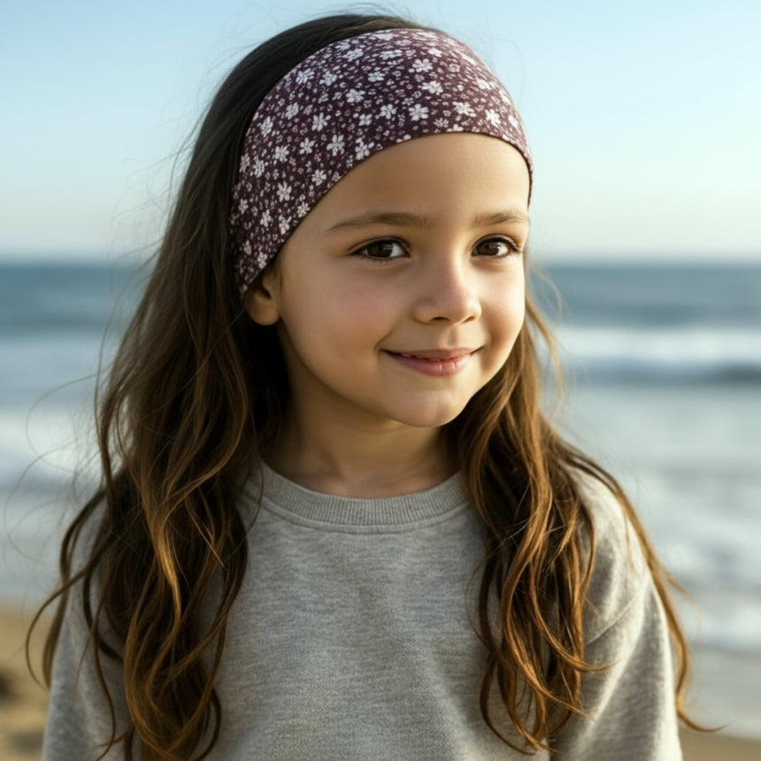 Ein Mädchen am Strand trägt ein Wollhuhn Elastisches Öko Stirnband, Jersey zweilagig, in Grau mit Blumenmuster, ideal für Outdoor-Abenteuer.