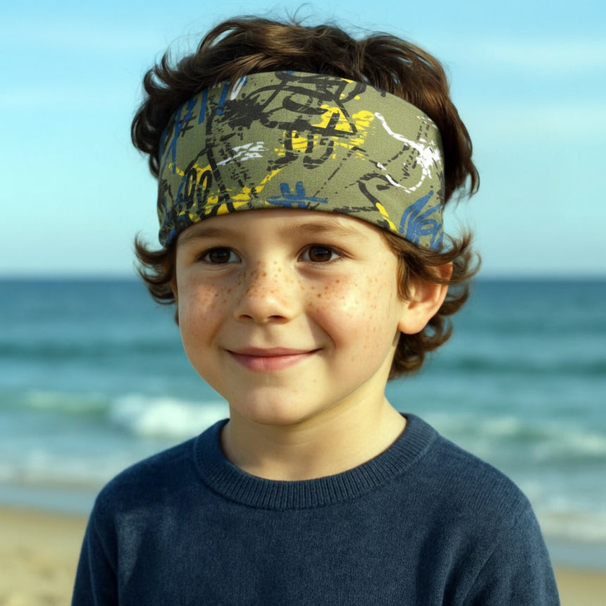 Junge mit Wollhuhn Elastisches Öko Stirnband, Jersey, zweilagig, breit, Graffiti-Design in Khaki/Gelb, auf Strand stehend, trägt blaues Sweatshirt. Ideal für Outdoor-Aktivitäten.