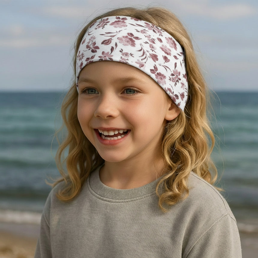 Mädchen am Strand trägt ein Wollhuhn Elastisches Öko Stirnband, Jersey, zweilagig, breit, mit Blumenmuster in Weiß/Altrosa, handgemacht und ideal für Outdoor-Aktivitäten.