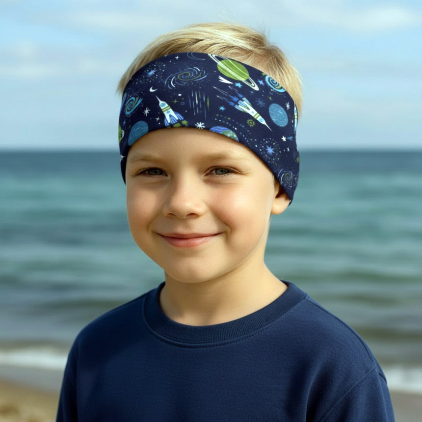 Kind am Strand trägt Wollhuhn Elastisches Öko Stirnband / Haarband Jersey Zweilagig Breit Space/Rakete Dunkelblau, lächelt sanft, Meer und Himmel im Hintergrund.