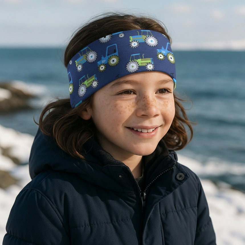 Kind in navyblauer Winterjacke trägt Wollhuhn Elastisches Öko Stirnband Jersey Zweilagig Breit Traktor/Trecker Dunkelblau, lächelnd am verschneiten Ufer stehend.