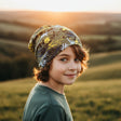 Kind mit lockigem braunem Haar trägt eine Wollhuhn Öko Long Beanie / Wendemütze in Graffiti Khaki/Gelb, steht lächelnd in einem Feld bei Sonnenuntergang.