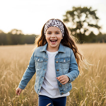 Mädchen mit Wollhuhn Elastisches Öko Stirnband Jersey Zweilagig Breit Leo Creme, läuft durch ein sonnenbeschienenes Feld, trägt Jeansjacke, weißes Shirt, dunkle Leggings.