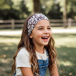Mädchen lacht im Freien, trägt Wollhuhn Elastisches Öko Stirnband Jersey Zweilagig Breit Leo Creme über weißem T-Shirt und Denim-Latzhose, ideal für Outdoor-Aktivitäten.