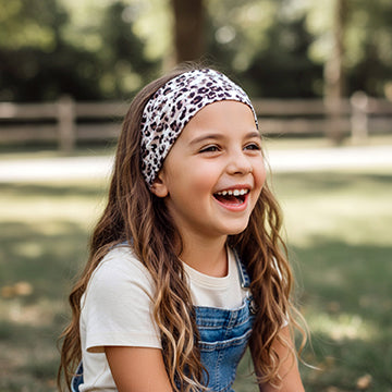 Mädchen lacht im Freien, trägt Wollhuhn Elastisches Öko Stirnband Jersey Zweilagig Breit Leo Creme über weißem T-Shirt und Denim-Latzhose, ideal für Outdoor-Aktivitäten.