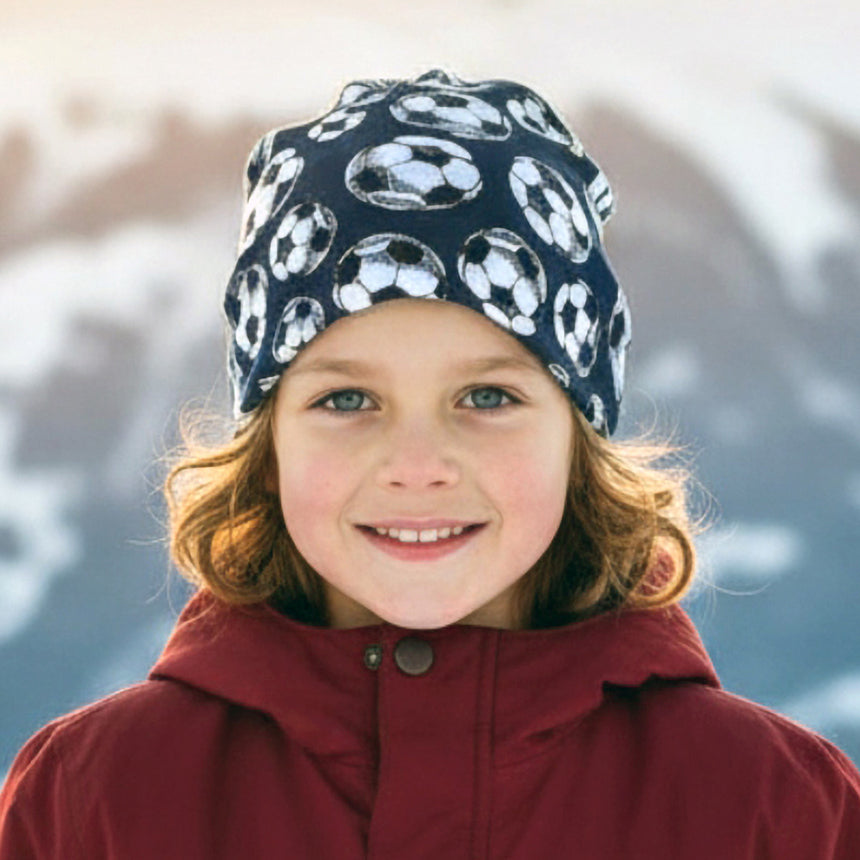 Kind im Schnee trägt die Wollhuhn Öko Long Beanie/Wendemütze in Dunkelblau mit Fußballmotiv. Handgemacht, zweilagig, perfekt für milde Wintertage.